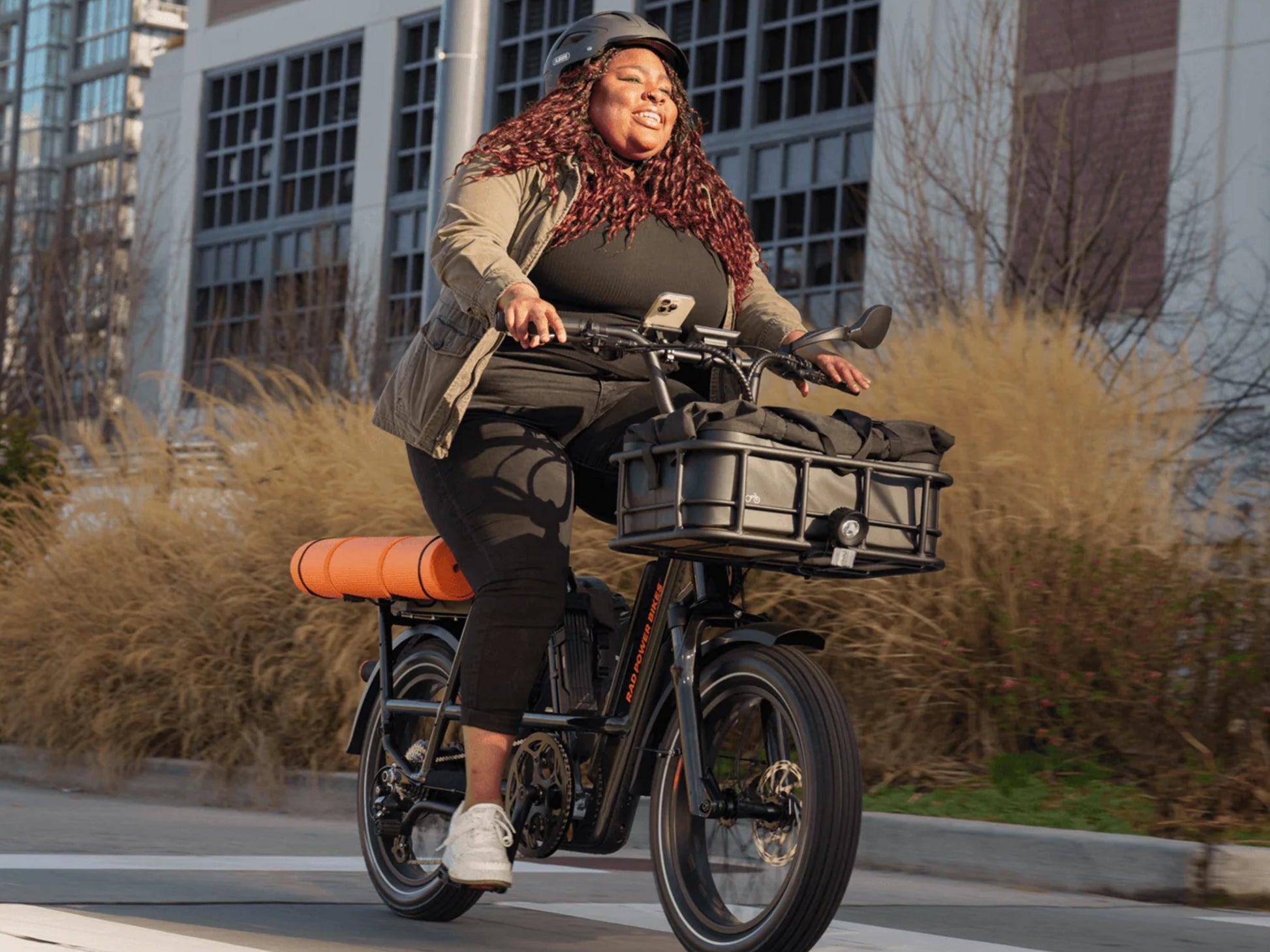 Rad Power RadRunner Max Electric Cargo Utility Bike. Woman riding on a paved street with a corporate building in background.