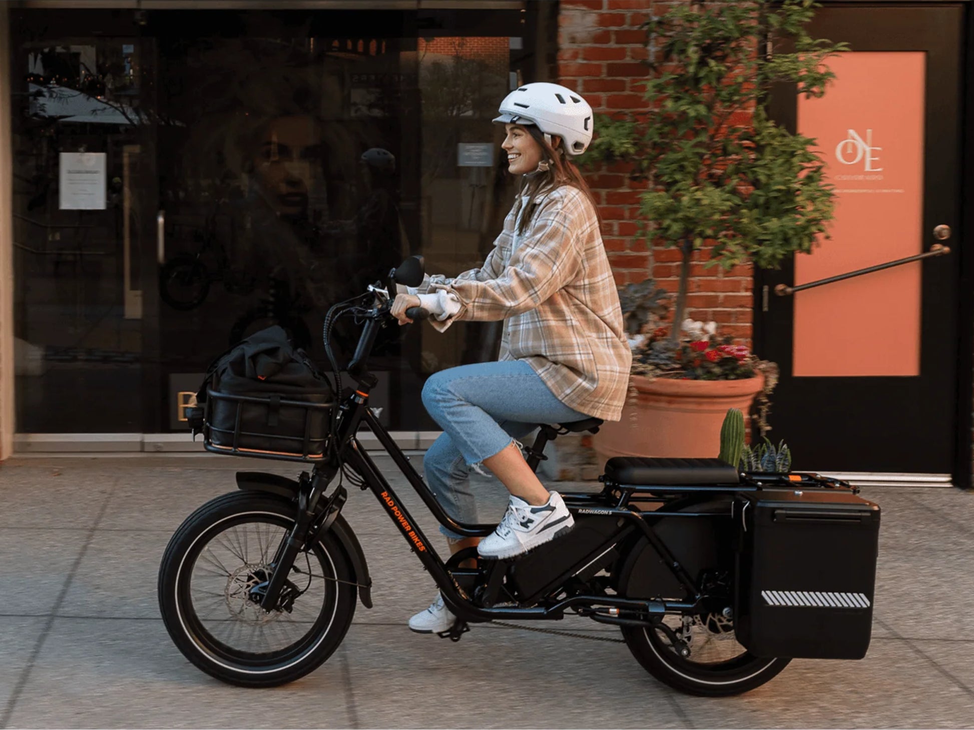Rad Power RadWagon 5 Electric Cargo bike Metalllic blue. Woman riding along shopping center. Optional accessories shown
