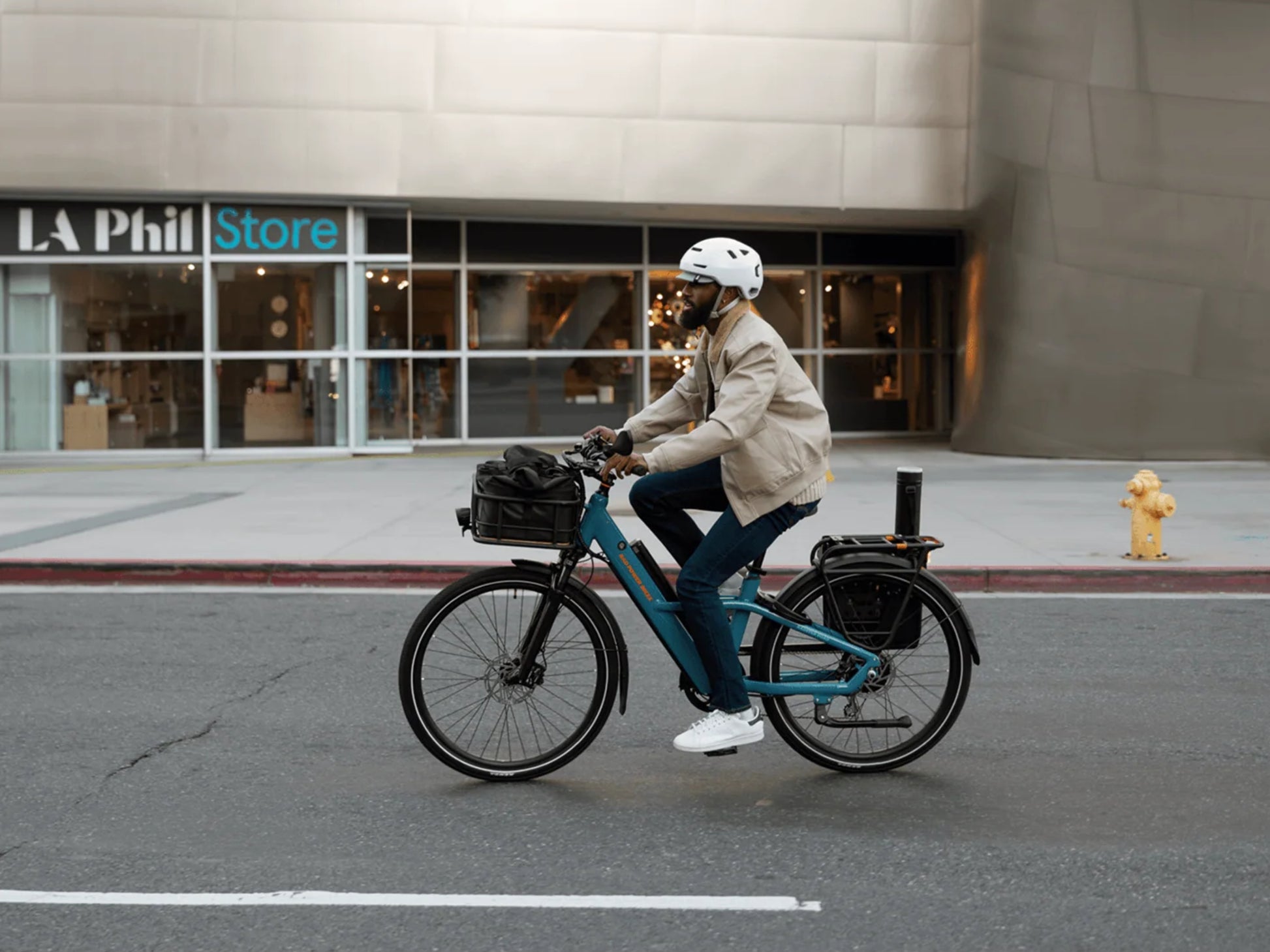 Rad Power Radster™ Road Electric Commuter Bike Bay blue. Man riding on the street  in front of a store with optional accessories shown
