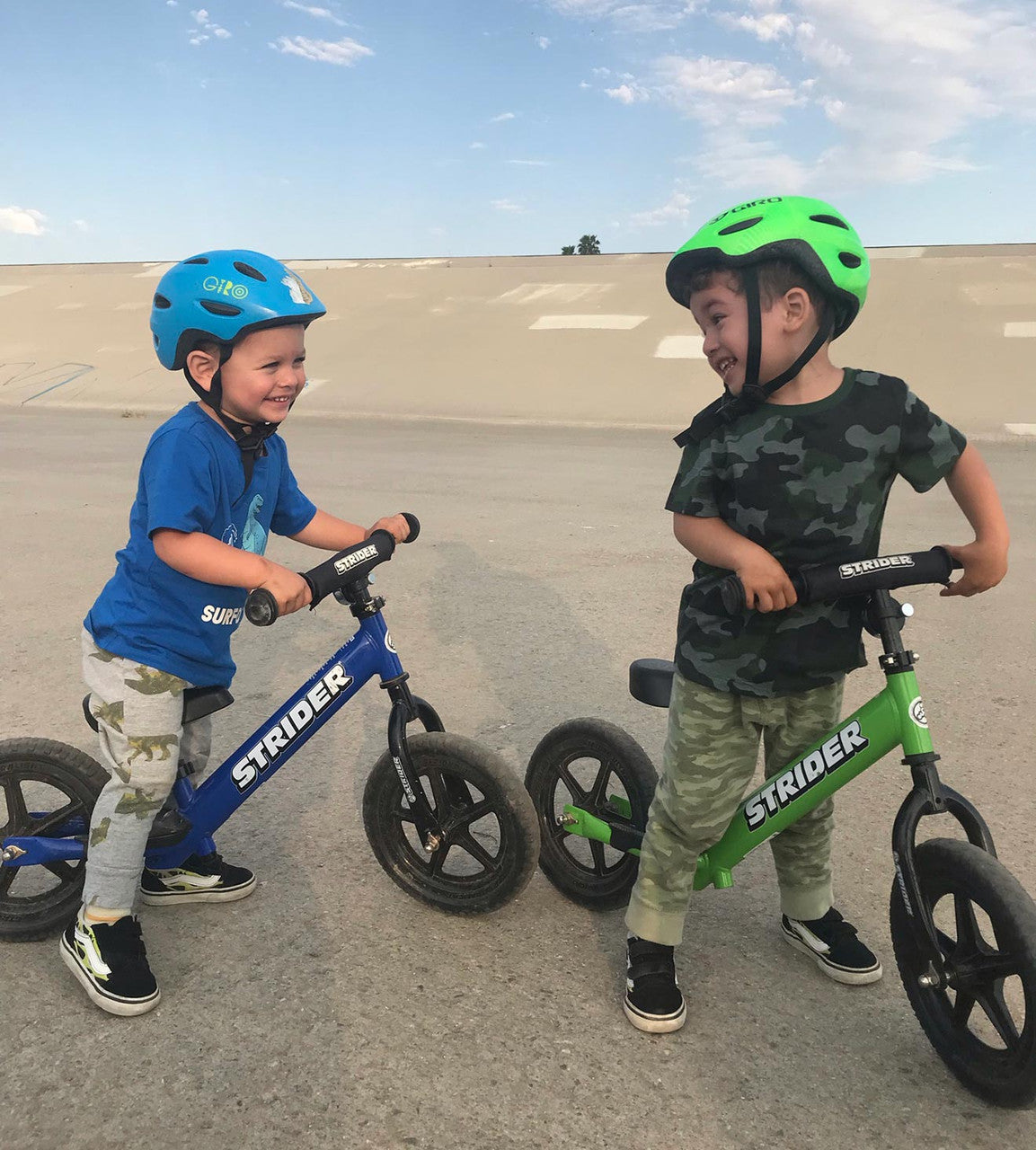 Strider 12 Balance Bike Sport Two children on green and blue bikes stopped and smiling on a path