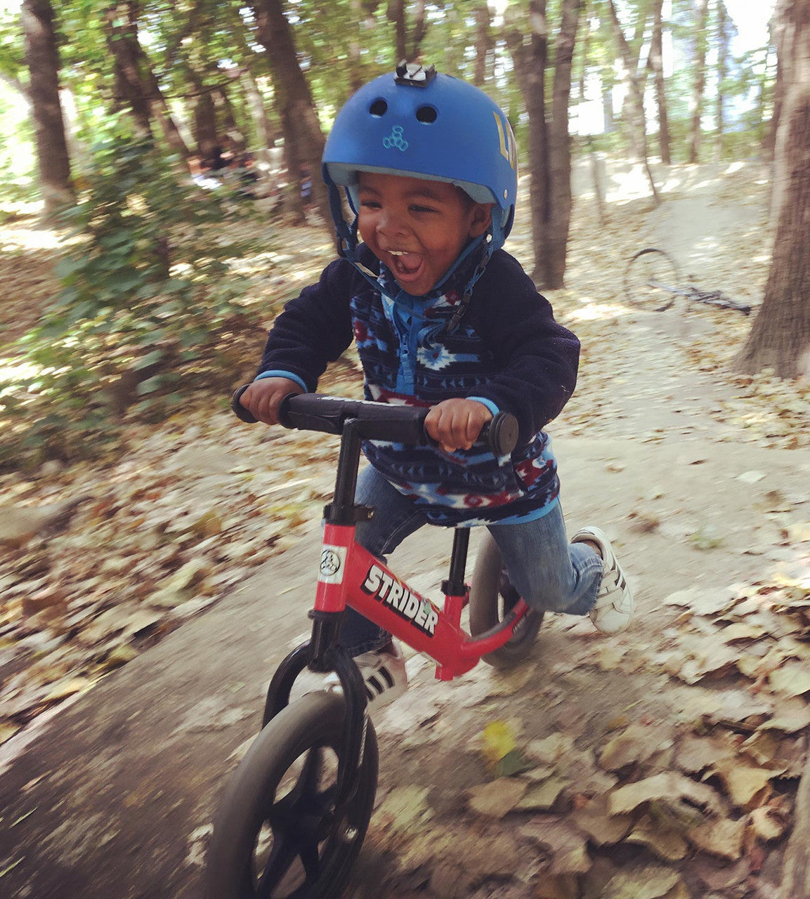 Strider 12 Balance Bike Sport Red Child with huge smile and blue helmet riding on dirt trail