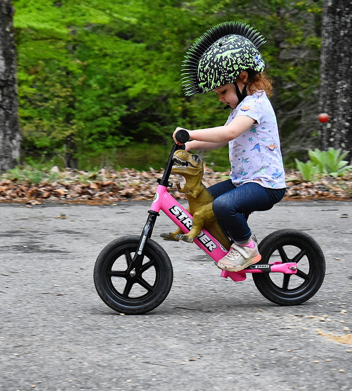 Strider Balance 12 Sport Pink Child riding with a dino spiked helmet and giving a dino toy a ride