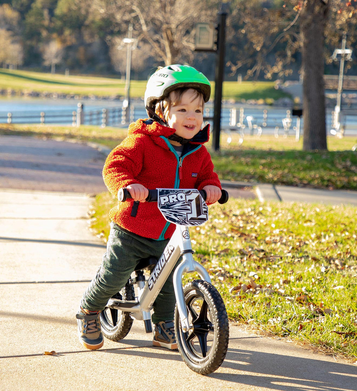 Strider 12 Pro Balance bike silver Child with a green helmet smiling on paved bike path with a lake in background