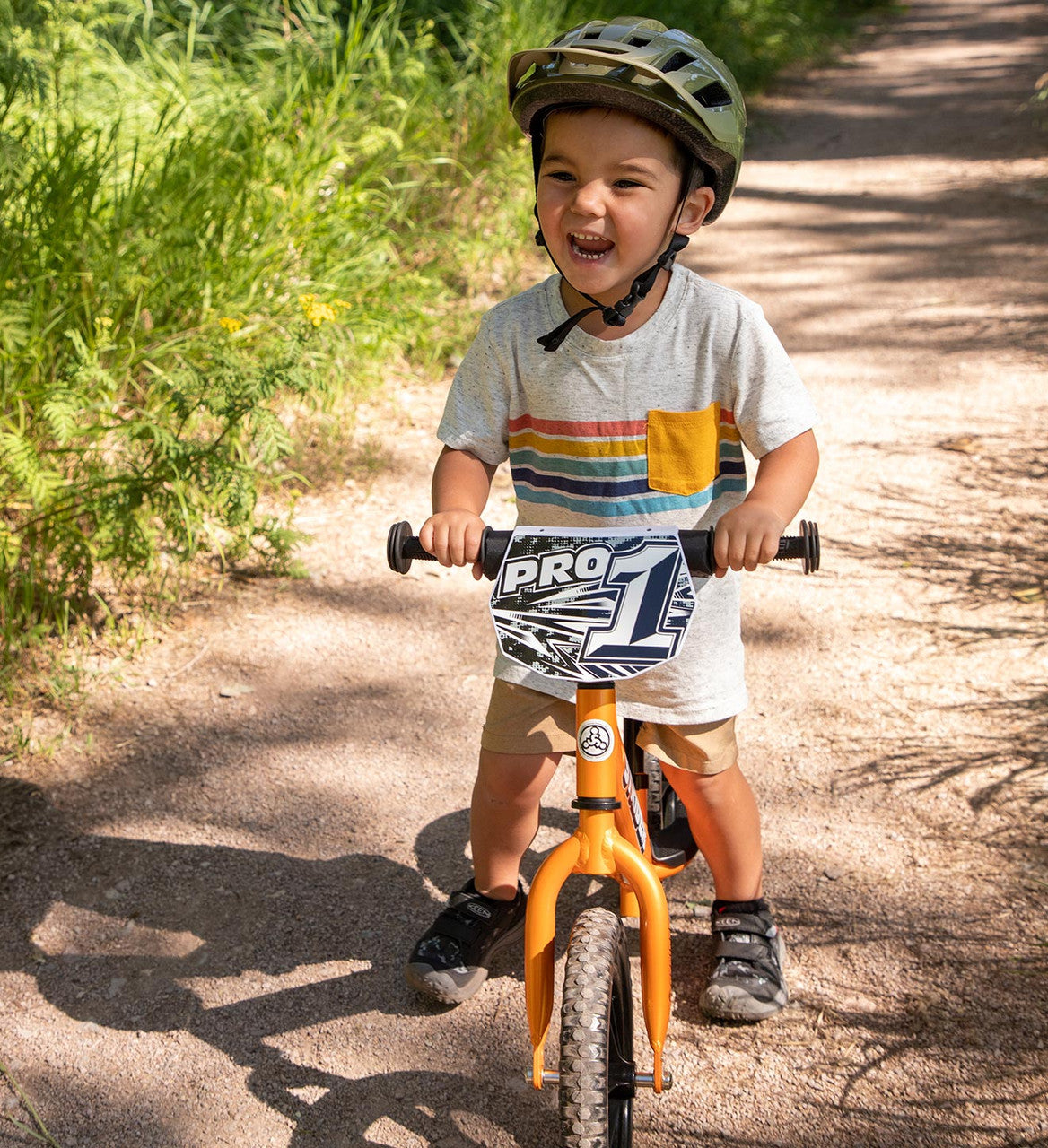 Strider 12 Pro Balance bike Orange rush Child with a huge smile and green helmet on striding on a dirt path